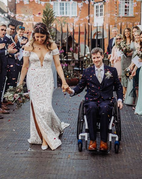 Bride in fitted wedding dress has confetti exit with groom in a blue suit in a wheelchair.