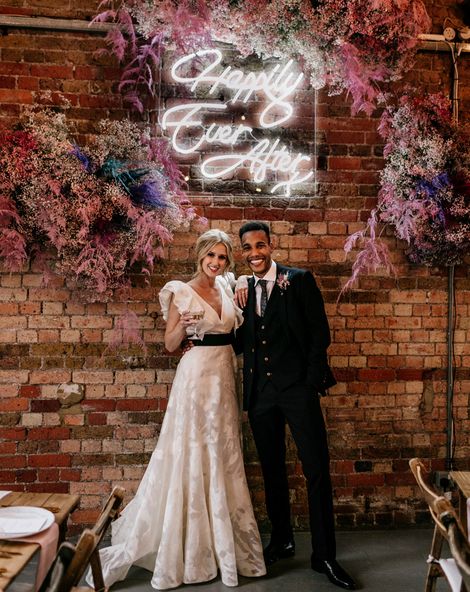 Prezola wedding couple standing in front of a neon happily ever after sign surround by pink gypsophila and wearing a ruffle sleeve wedding dress