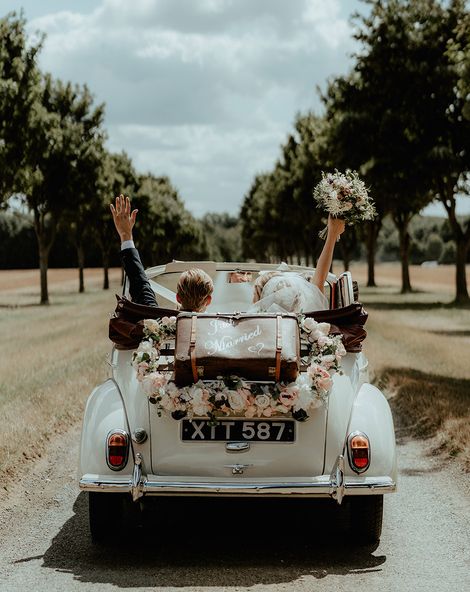 Bride and groom ride away in their wedding car with an old fashioned suitcase and flower decor.