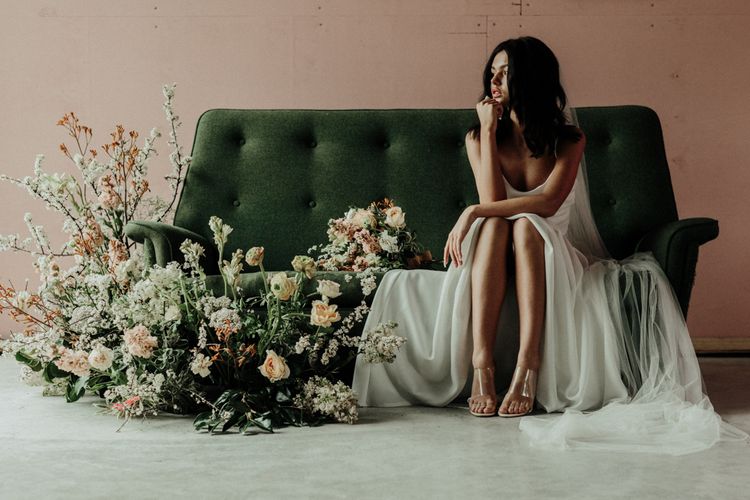 Bride in tulle wedding dress sitting on a forest green sofa with floral arrangement