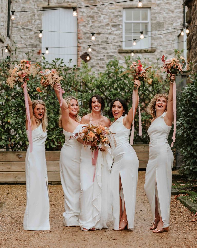 Bride in Alexandra Grecco dress with bridesmaids in white one shoulder dresses and neutral wedding bouquets.